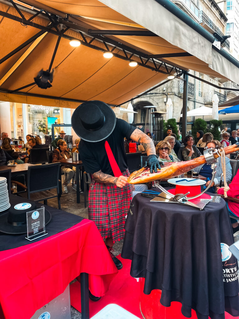 Galician Ham Carver realizando corte artístico de jamón ibérico durante una colaboración, ofreciendo una experiencia gastronómica en vivo para los asistentes en una terraza