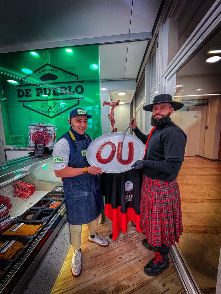 Cortador de jamón profesional en Ourense junto a carnicero local en Carnicería De Pueblo, mostrando bandeja con letras OU hechas con lonchas de jamón ibérico.