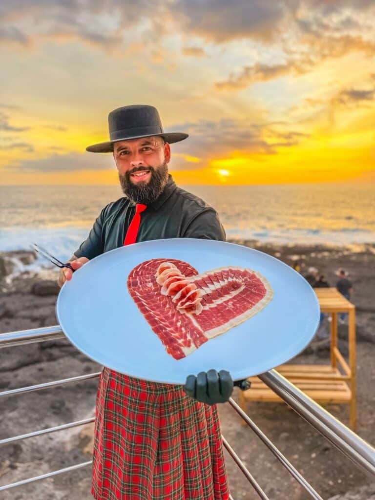 Plato de jamón ibérico en forma de corazón al atardecer – Galician Ham Carver Playa Paraíso Julio García, Galician Ham Carver, sosteniendo un plato de jamón ibérico en forma de corazón al atardecer en Playa Paraíso, Tenerife Sur